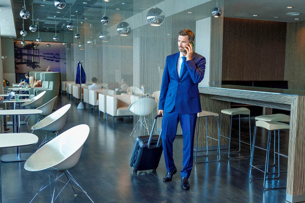 Elegantní muž v modrém obleku s cestovním kufrem telefonuje v moderním prostředí Erste Premier Lounge.