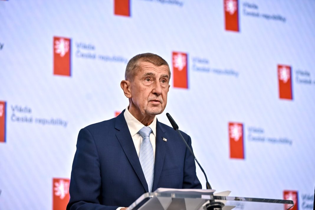 Muž v tmavém obleku hovoří u řečnického pultu před logy Vlády České republiky