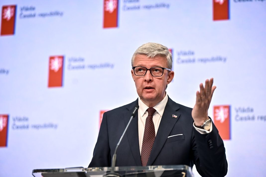 Karel Havlíček hovoří u řečnického pultu před pozadím s logy vlády České republiky